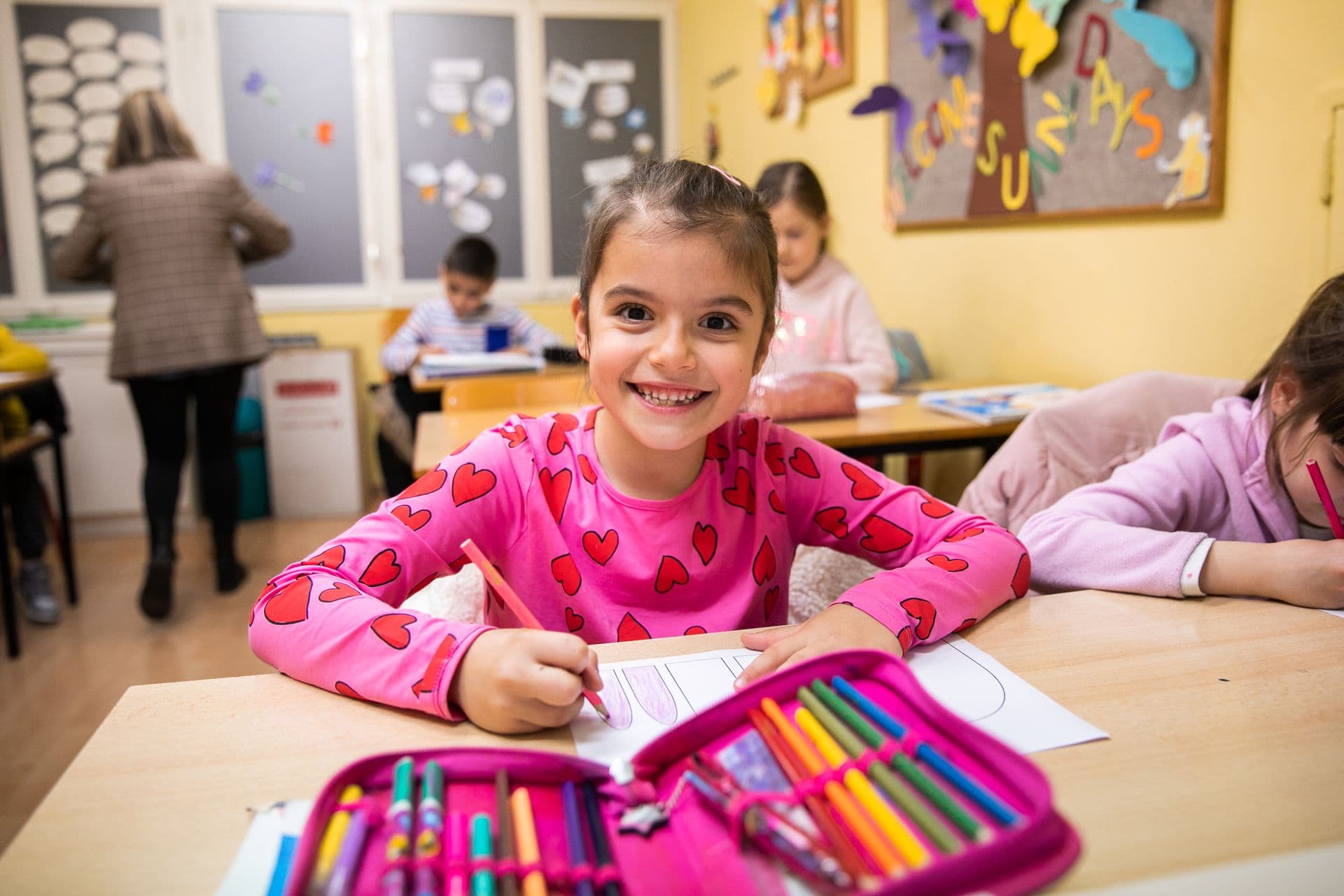 A smiling girl at a school desk with coloured pencils